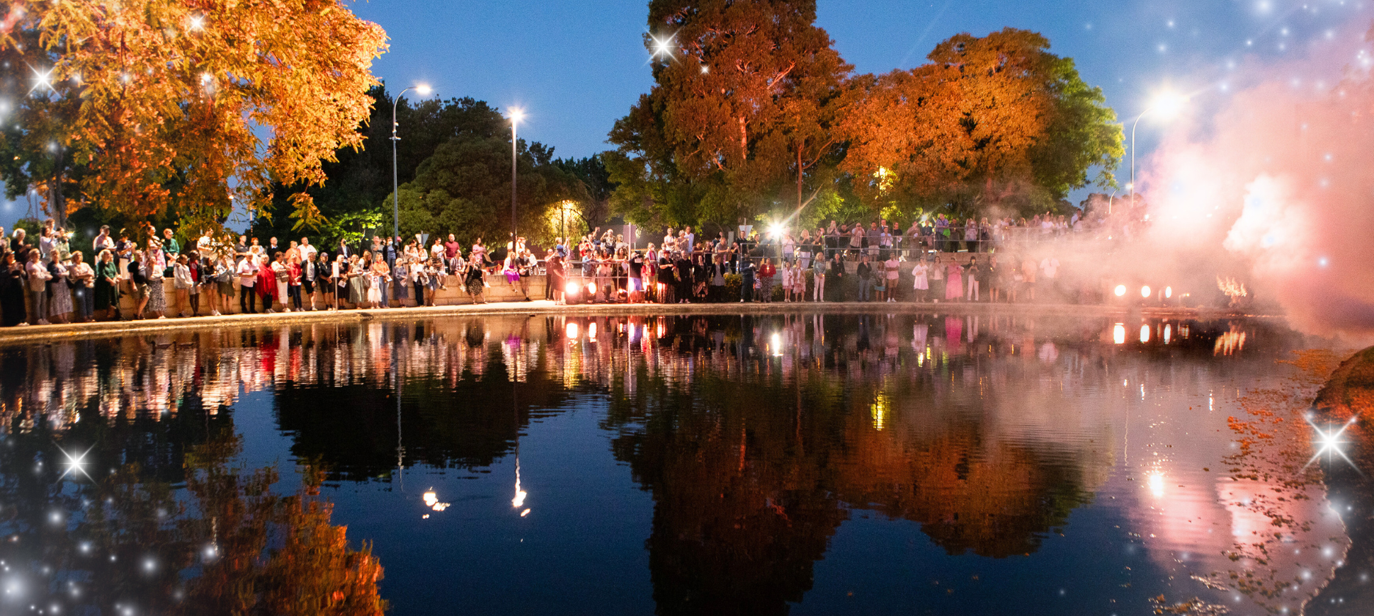 Image of a large group of people standing by the edge of a lake at St John of God Murdoch Hospital. It is night time, and there are lights twinkling across the lake and in the sky. 