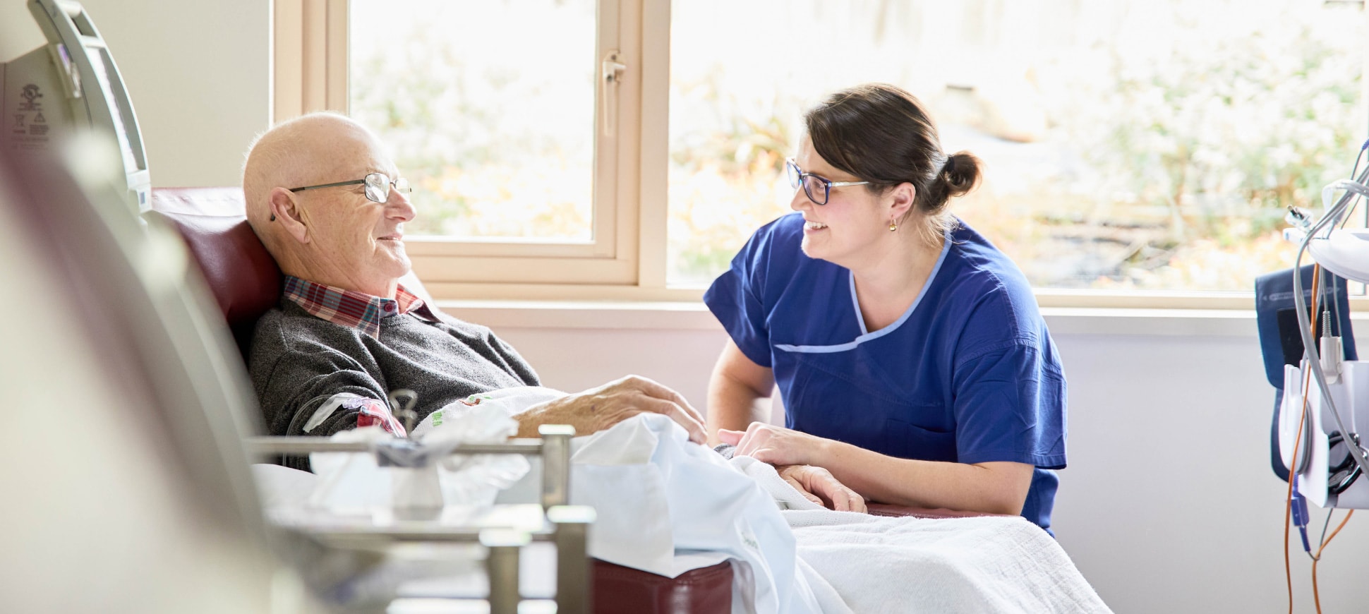 A caregiver bends down to talk to an oncology patient reclining in a chair