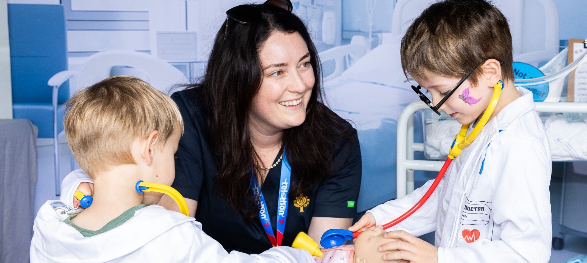 Caregiver interacts with two young boys dressed up in doctors white coats and plastic stethoscopes