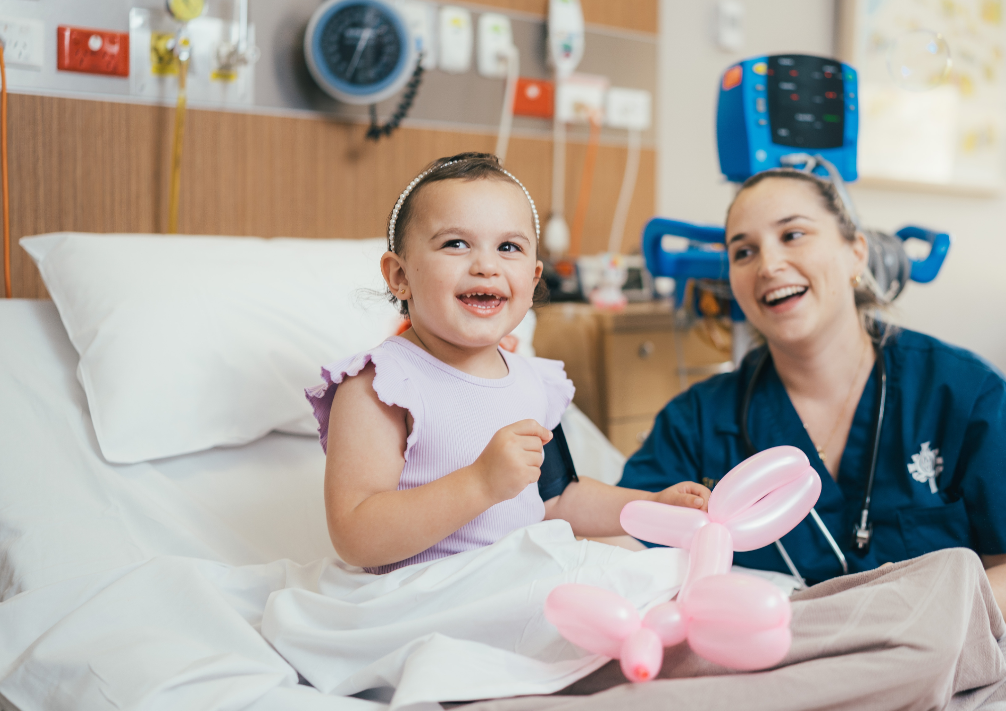 Caregiver and young patient sitting on hospital bed and smiling. The young patient is holding a balloon/ 