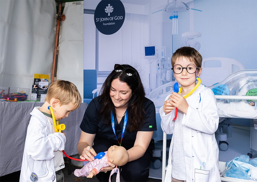 A caregiver holding a toy stethoscope to a doll as two young children dressed as doctors look on.