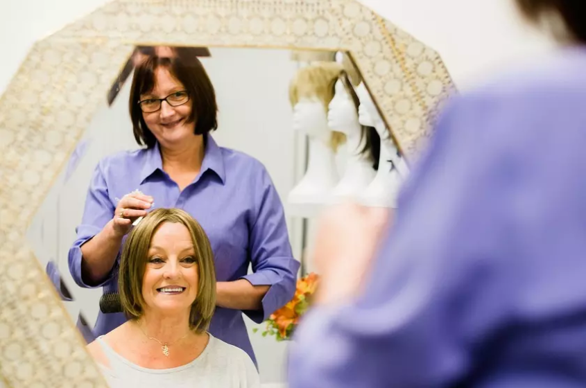 Caregiver helping patient put on wig, both are looking into a mirror and smiling. 